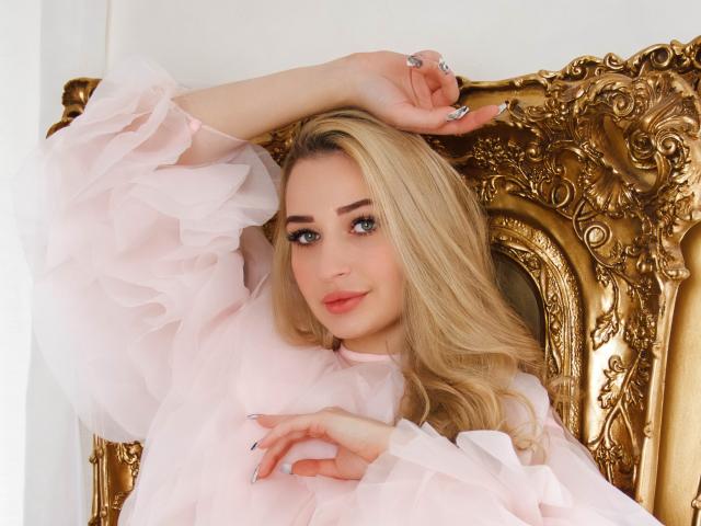 Barbara Rosy, mit ihren langen blonden Haaren, lehnt sich in einem verzierten goldfarbenen Stuhl zurück und trägt ein durchsichtiges rosa Oberteil. Ihre Hand ruht auf ihrem Kopf und strahlt eine selbstbewusste, verführerische Ausstrahlung aus. Helle Beleuchtung betont ihre Gesichtszüge..