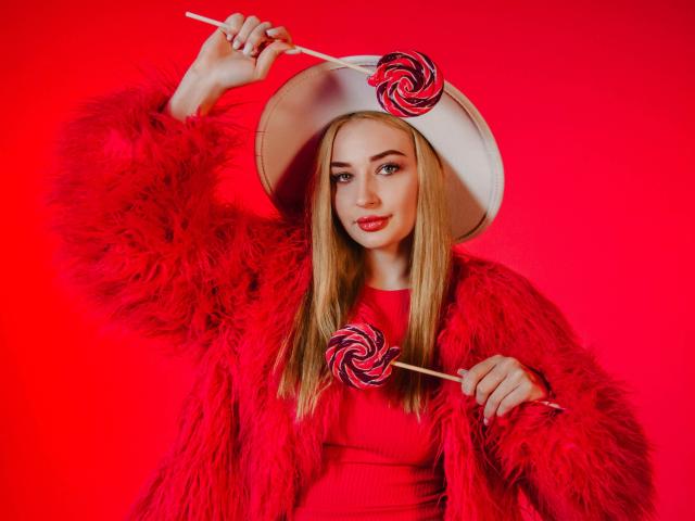 Barbara Rosy, avec de longs cheveux blonds, des lèvres rouges, portant un manteau en fourrure rouge et un chapeau blanc, tenant deux sucettes devant... Fond rouge vif. Son expression est sensuelle et séduisante..