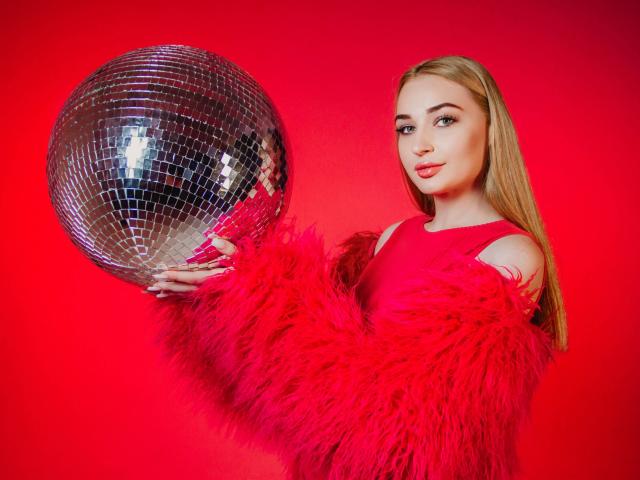 BarbaraRosy segura uma brilhante bola de discoteca prateada contra um fundo vermelho vibrante. Ela veste uma blusa vermelha, peluda, com um ombro descoberto, e tem longos cabelos loiros. Visual elegante, confiante e sensual..