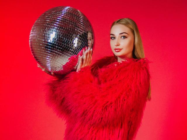 Barbara Rosy, een blonde vrouw met rode lippenstift, houdt een glanzende zilveren discobal vast voor een felrode achtergrond. Ze draagt een rode, pluizige jas en straalt een zelfverzekerde, aantrekkelijke uitstraling uit..