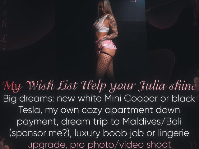 LexiLee, una mujer rubia con tatuajes, está de pie en una habitación oscura, vestida con un top de lencería rojo y medias de red. Ella sostiene un letrero de neón brillante, creando un ambiente sensual y provocador..
