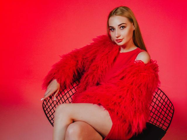 Barbara Rosy está sentada en una silla de alambre negra, con las piernas cruzadas, y lleva un abrigo rojo de piel sintética esponjosa que deja al descubierto sus hombros. muslos. Fondo rojo, iluminación roja intensa, expresión sensual..