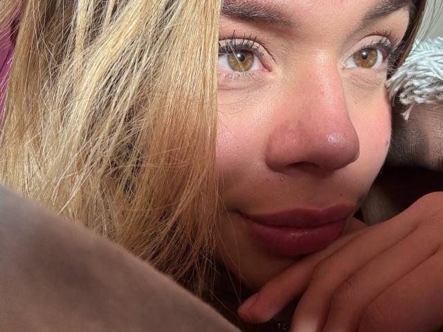 Foto em close-up de EmmaRiveraa, uma mulher de pele clara, com olhos castanhos, lábios ligeiramente entreabertos e cabelo loiro, parcialmente visível. Cobrir o rosto. A mão dela repousa sob o queixo, criando uma atmosfera sensual e íntima..