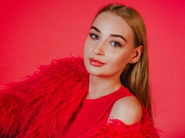 BarbaraRosy with long blonde hair, wearing a red dress and a fluffy red fur coat, against a bright red background. She has red lipstick and a sultry expression. Fucking hot.