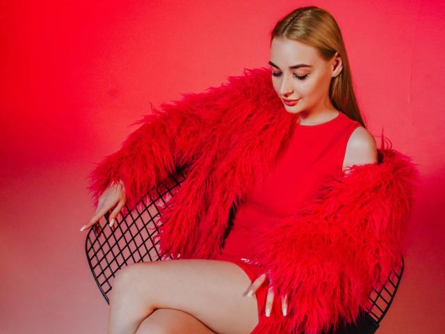 Barbara Rosy est assise sur une chaise avec un dossier en mesh noir, portant un manteau en plumes rouges et une robe rouge assortie. Elle regarde vers le bas, les jambes croisées, sur un fond dégradé rouge et gris. Extrêmement sexy..