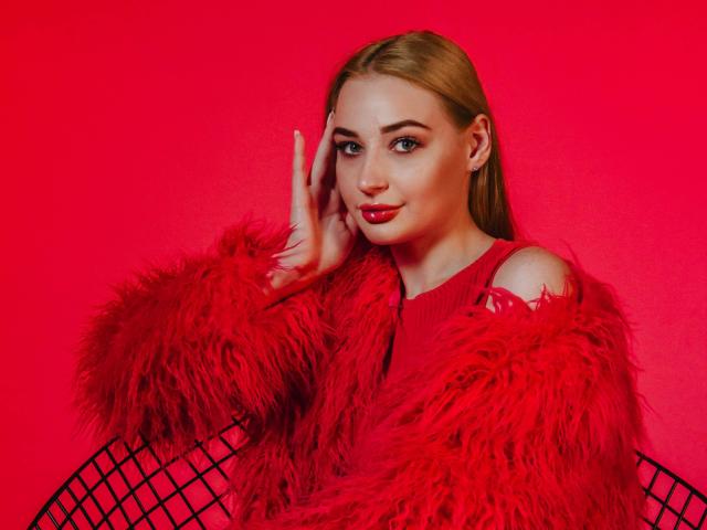Barbara Rosy, met glad achterover gekamd blond haar, draagt een rode bontjas, een rode jurk en een stoel met netstof. Ze heeft haar hand op haar wang, en alles is rood. lippen, intense blik, rode achtergrond, levendig, sexy, gedurfd.