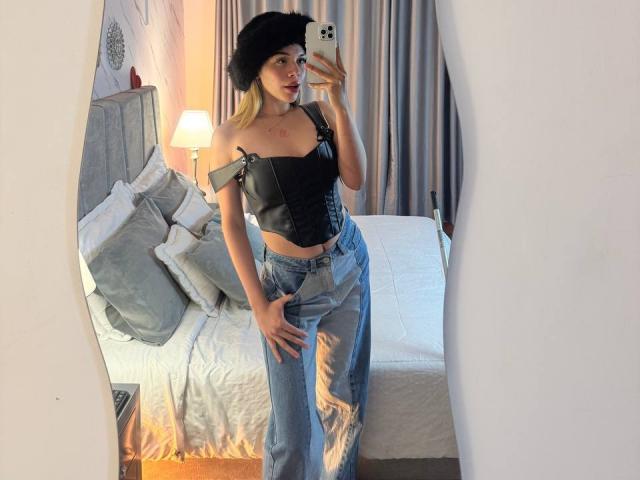 EmmaRiveraa, sexy as fuck, standing in bedroom, taking mirror selfie, wearing black off-shoulder top, ripped jeans, black beret, holding smartphone, light from lamp casting glow, bed with pillows in background.