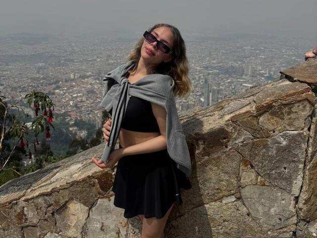 EmmaRiveraa leunt tegen een stenen muur, ze draagt een zwarte crop top en rok, met een grijze sjaal om haar schouders. Ze draagt een zonnebril op en kijkt verleidelijk om. Stadsgezicht op de achtergrond.