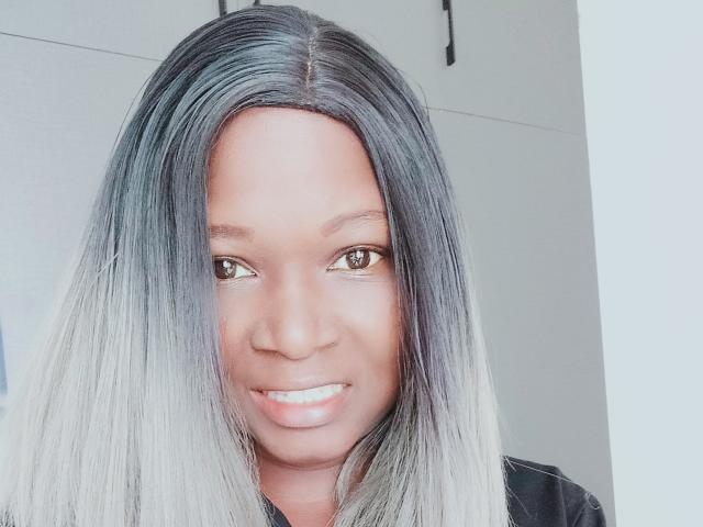 PrettyAriah with long, straight, gray-to-black ombre hair, smiling, brown eyes, and smooth dark brown skin, wearing a black shirt. She's looking at the camera with a gentle smile.
