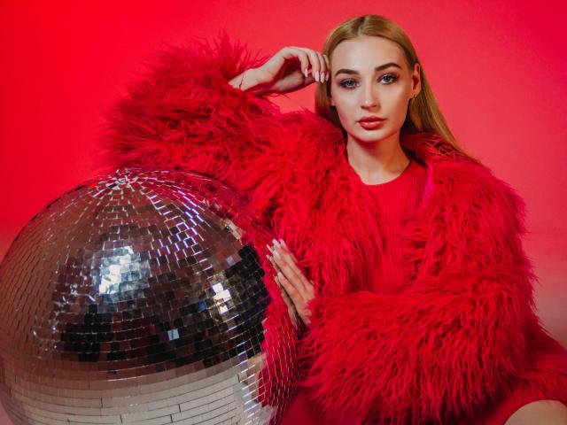 BarbaraRosy, sexy, sits next to a shiny silver disco ball, wearing a bright red, fluffy fur coat. Red background. She's looking directly at the camera, exuding confident sex appeal.