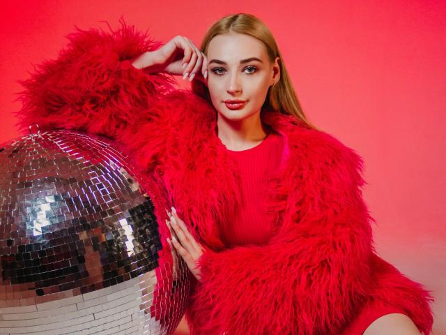 BarbaraRosy in a bright red, fluffy fur coat, leaning on a shiny silver disco ball against a gradient red background. She's smirking, with one hand in her hair, exuding confidence and sexiness.