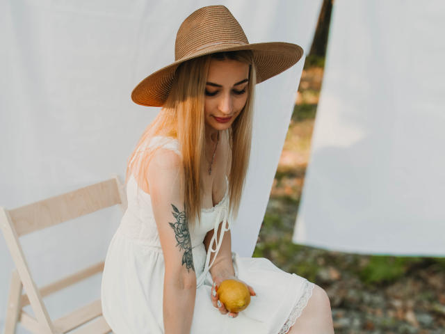 Barbara Rosy está sentada em uma cadeira, vestindo um vestido branco e um chapéu de palha, segurando uma laranja. Seus longos cabelos loiros caem sobre seus ombros, e uma tatuagem é visível em seu braço esquerdo. A luz do sol realça seu rosto..