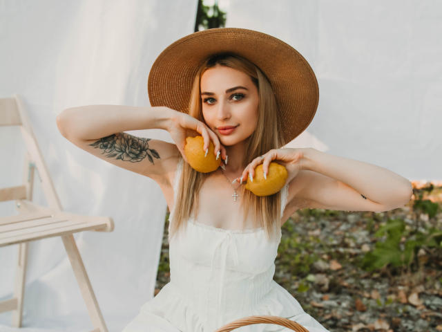 BarbaraRosy holds two orange peaches, wearing a white dress and straw hat, with a floral tattoo on her left arm. Her long hair is blonde, and she's smiling slightly. Background includes a white ladder and greenery.