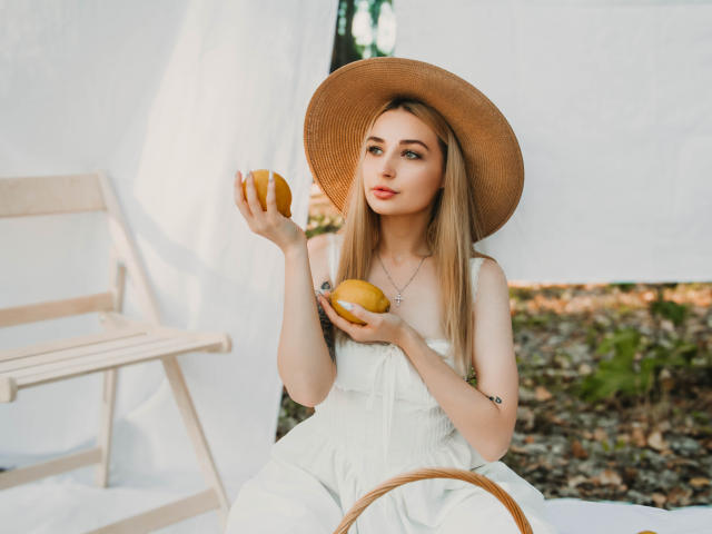 Barbara Rosy, una mujer rubia atractiva con cabello largo, lleva un sombrero de paja de ala ancha y un vestido blanco, sosteniendo un limón amarillo. En su mano derecha, mirada intensa, entorno exterior luminoso..