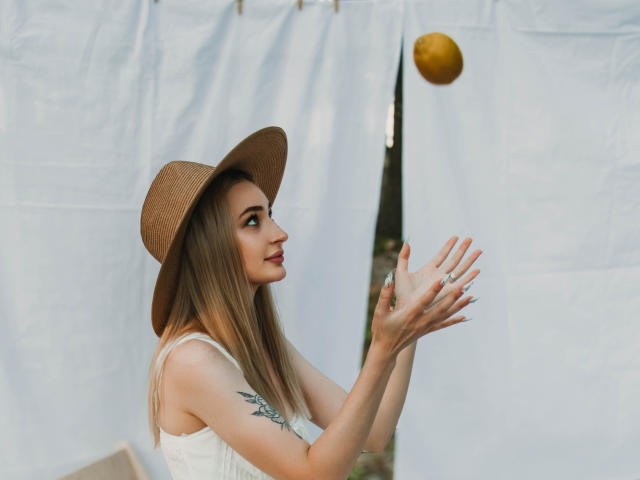 BarbaraRosy, fair-skinned woman with long brown hair, wearing a brown hat and white sleeveless top, has a tattoo on her upper arm. She's catching a ball, with white cloth background. Fucking intense focus.