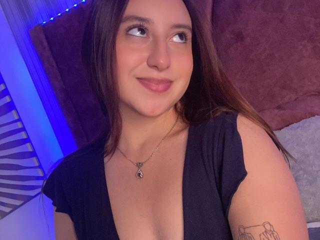 RosalieDupont with long brown hair, smiling slightly, wearing a black top with a low neckline, necklace with a gem, small cat tattoo on left arm, looking to the side. Fucking sexy.