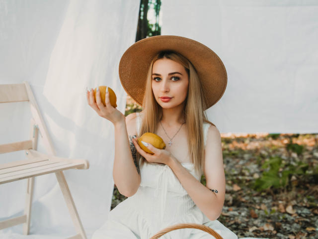 BarbaraRosy sits holding an orange, wearing a white dress and wide-brimmed hat, with a second orange in her lap. Her long blonde hair, red lips, and seductive gaze dominate.