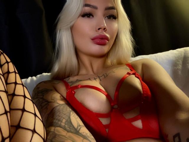 LexiLee, una donna bionda con tatuaggi, indossa un reggiseno di pizzo rosso con aperture, calze a rete, labbra tumefatte, sguardo seducente. illuminazione soffusa, sfondo scuro, forte tensione sessuale, primo piano, posa provocante.