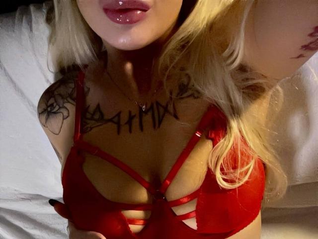 Foto de primer plano de LexiLee con cabello rubio, un sujetador rojo con tirantes, labios brillantes, tatuajes visibles en el pecho, y una expresión seductora. expresión, fuerte tensión sexual, escote pronunciado, contrastes de luz y sombra.
