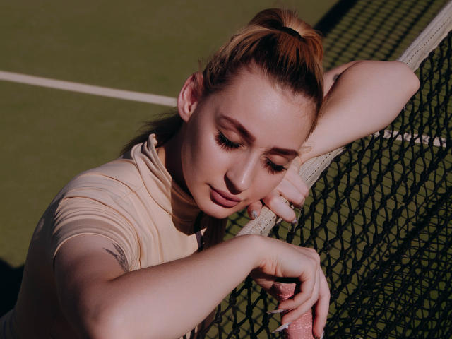 BarbaraRosy, a young woman with fair skin and brown hair in a ponytail, leans against a tennis net, eyes closed, wearing a beige shirt. Sunlight highlights her relaxed, sexy pose.