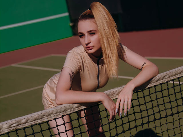 BarbaraRosy with a ponytail, leaning over a tennis net, wearing a tight, beige dress, looking directly at the camera, tattoo on left arm, sexy, intense gaze, athletic build.