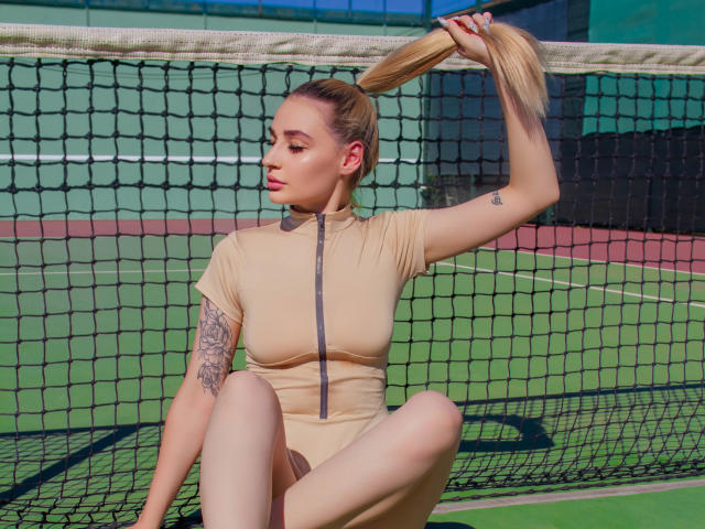 Barbara Rosy zit op een tennisbaan en houdt haar paardenstaart met één hand vast. Ze draagt een strak, beige body met rits, waardoor een tatoeage op haar linkerarm zichtbaar is. Helder zonlicht. Verdomd aantrekkelijk..