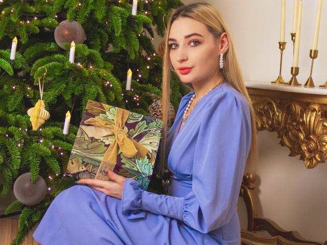 Barbara Rosy está sentada frente a un árbol de Navidad con velas encendidas, vestida con un vestido azul y sosteniendo un regalo envuelto de colores. regalo. Tiene lápiz labial rojo, cabello rubio y aretes colgantes. ¡Qué atractiva!.