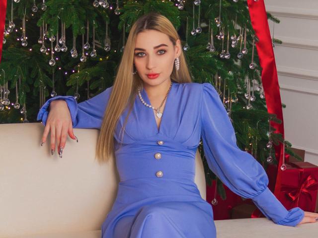 BarbaraRosy, a blonde woman with long hair, sits on a white couch, wearing a blue dress with buttons, looking directly at the camera. Christmas tree with ornaments and red ribbons in the background. Sexual tension.