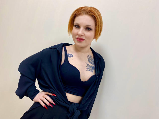 Foto van Selinn, een vrouw met bleke huid en kort rood haar, die een zwarte bh en een open zwart shirt draagt, met haar hand op haar heup. Rode nagels, tatoeage op de borst, grijns, witte achtergrond..