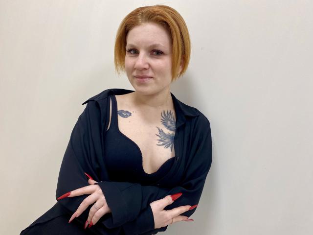 Photographie de Selinn, une femme rousse à la peau pâle, portant un soutien-gorge noir visible sous une chemise noire ouverte. Elle a des tatouages sur sa poitrine, les bras croisés, et elle est légèrement penchée en avant..