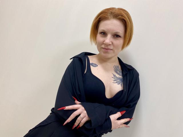 Fotografie von Selinn, einer hellhäutigen Frau mit kurzen, roten Haaren, die ein schwarzes Kleid mit tiefem Ausschnitt und Ärmeln trägt. überkreuzte Arme, Tätowierung auf der Brust sichtbar, rote Fingernägel, weißer Hintergrund, sexy Ausstrahlung..