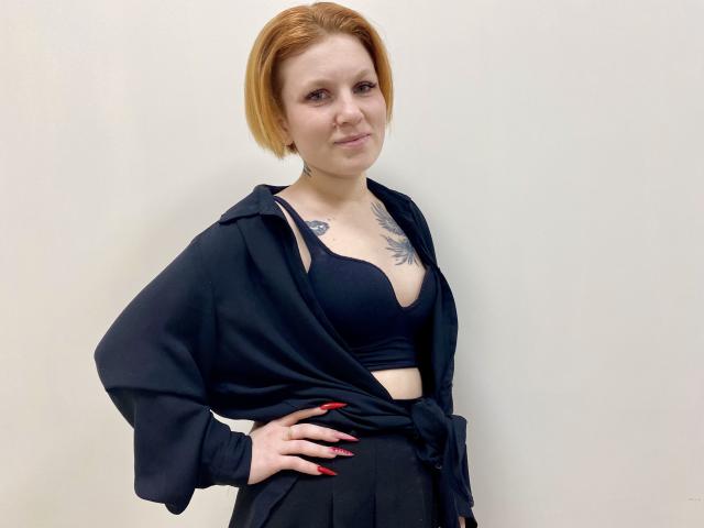 Photographie de Selinn, une femme à la peau claire avec des cheveux courts roux, portant un soutien-gorge noir et une robe noire ouverte, les mains sur les hanches. Tatouage sur la poitrine, vernis à ongles rouge, pose confiante..