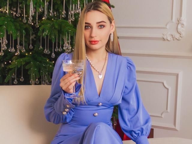 Barbara Rosy, met lang blond haar, een blauwe jurk met pofmouwen, die een martini-glas vasthoudt, zittend, sexy, zelfverzekerd. rode lippenstift, witte muur, groene kerstboom, glinsterende versieringen, verleidelijke blik.