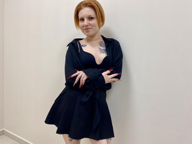 Photographie de Selinn, debout devant un mur blanc, les bras croisés, portant une robe noire avec un décolleté plongeant, laissant entrevoir des tatouages sur sa poitrine et ses bras, des cheveux roux, une expression confiante.