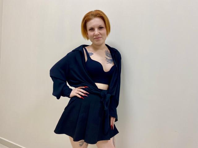 Fotografia de Selinn, em pé contra uma parede branca, vestindo uma blusa cropped e uma saia pretas, com uma camisa preta amarrada, vermelha cabelo, tatuagens visíveis no peito e no braço, mão na cintura, expressão confiante..