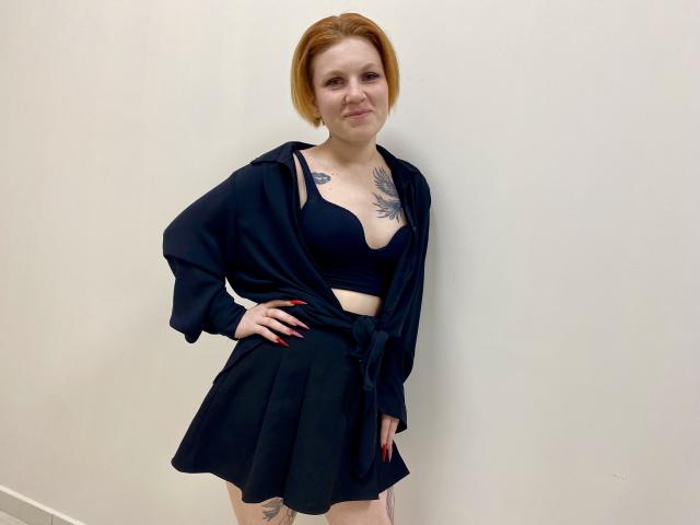 Fotografia de Selinn, em pé contra uma parede branca, vestindo um sutiã preto e uma camisa preta amarrada, uma saia preta e uma bermuda vermelha. cabelo, tatuagens no peito, mão na cintura, sorriso confiante, pose sensual..