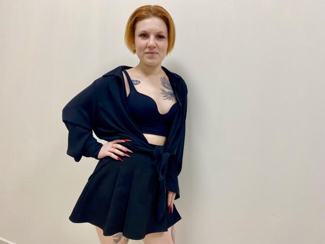 Photographie de Selinn, debout devant un mur blanc, portant un soutien-gorge et une jupe noirs, avec une chemise noire nouée. Elle a les cheveux roux, des tatouages sur la poitrine et une posture assurée. Sa main repose sur sa hanche, avec un léger sourire sur son visage..