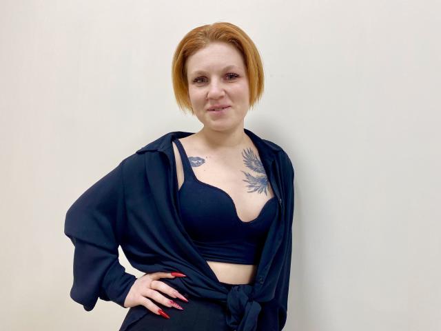 Photographie de Selinn, une femme à la peau claire avec des cheveux courts roux, portant un soutien-gorge noir et une chemise noire nouée, laissant entrevoir gros tatouage sur la poitrine, posé devant un fond blanc. Extrêmement sûr de soi..