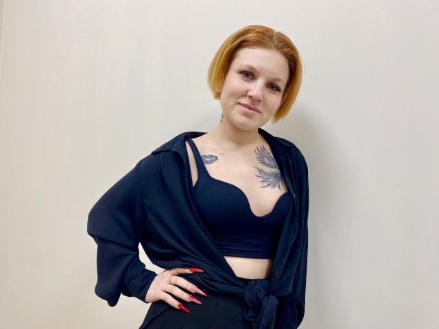 Photographie de Selinn, une femme à la peau claire avec des cheveux courts roux, portant un soutien-gorge en dentelle noire et un cardigan noir ouvert. exposant sa poitrine tatouée et son vernis à ongles rouge. Pose incroyablement séduisante..