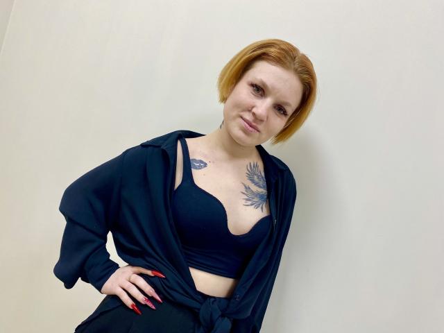 Fotografía de Selinn, una mujer de piel clara con cabello corto color naranja, que lleva un sujetador negro y una camisa negra abierta, mostrando un tatuaje en el pecho, de pie con la mano en la cadera, inclinándose hacia adelante.