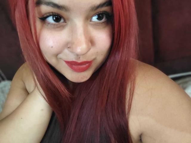 Close-up photo of AmberCutel with long red hair, brown eyes, and red lipstick. She has a nose piercing and freckles. AmberCutel's arm is raised, partially covering her bare shoulder. Sexual tension.