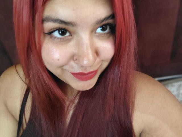 Close-up photo of AmberCutel with long red hair, dark eyebrows, black eyeliner, red lipstick, and a nose piercing. She's looking directly at the camera with a flirtatious smile, showing her cleavage.