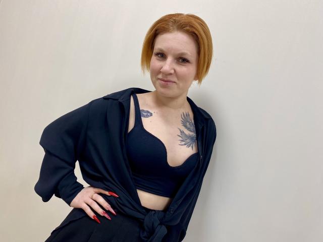 Fotografia de Selinn, uma mulher de pele clara com cabelo curto ruivo, vestindo um sutiã preto e uma camisa preta aberta, revelando tatuagem floral em seu peito. Mãos nos quadris, unhas vermelhas, sorriso discreto..