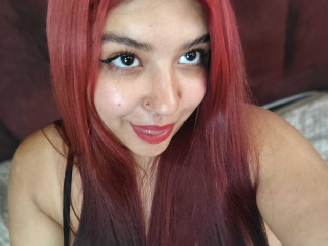 Close-up of AmberCutel with red hair, black eyeliner, and red lipstick, smiling at the camera. AmberCutel has a nose piercing and black tank top. Fucking hot, with intense eye contact.