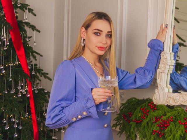 Barbara Rosy, con lunghi capelli biondi, indossa un abito blu con bottoni, tiene un bicchiere, appoggiata a uno specchio decorato, con un albero di Natale. un nastro rosso e degli ornamenti a sinistra, bacche rosse tra il verde sottostante. Molto attraente..