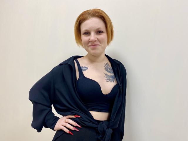 Photographie de Selinn, une femme à la peau claire avec des cheveux courts roux, portant un soutien-gorge noir et un cardigan noir ouvert, montrant une tatouage représentant un soleil éclatant sur sa poitrine. Main posé sur la hanche, expression confiante..