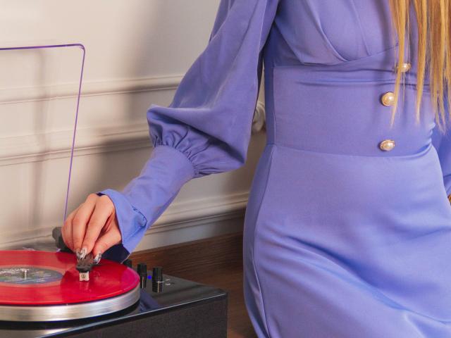 Barbara Rosy, vestida de azul con mangas abullonadas, sosteniendo con su mano derecha una aguja roja para tocadiscos, su cabello rubio largo cabello parcialmente visible. Tensión sexual proveniente de su agarre concentrado..