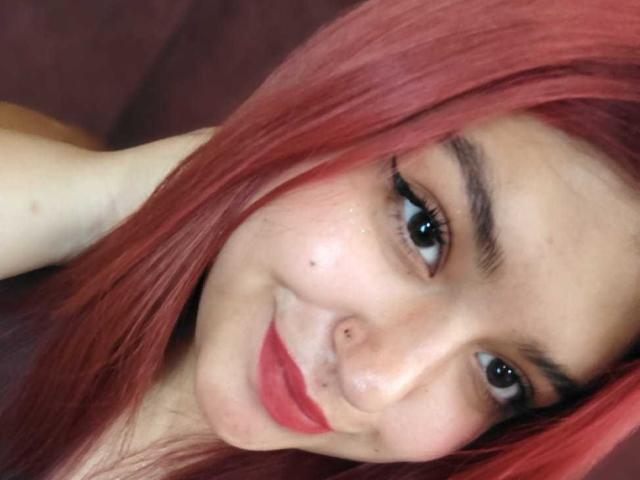 Foto em close-up de AmberCutel com cabelo ruivo, pele clara e delineador preto. Ela tem um piercing no nariz, batom vermelho e um sorriso discreto. Seu olhar é direto, transmitindo uma vibe sensual..