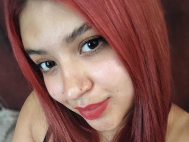 Close-up photo of AmberCutel with red hair, light skin, and black eyes, wearing red lipstick, looking directly at the camera with a slight smile, showing subtle facial freckles. Fucking sexy.