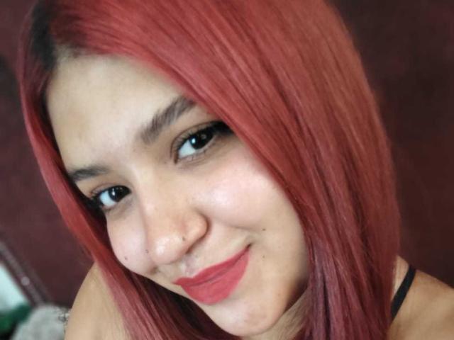 Photo en gros plan d'AmberCutel avec des cheveux rouges lisses, une peau claire et des yeux foncés, souriant légèrement avec du rouge à lèvres rouge. On peut voir son débardeur noir. Elle regarde directement l'appareil photo..